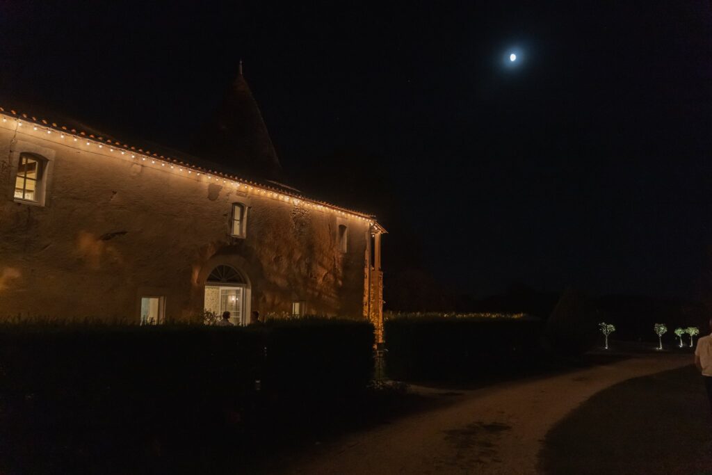 La façade en pierre du château éclairée de guirlandes, de nuit, avec la lune dans le ciel.