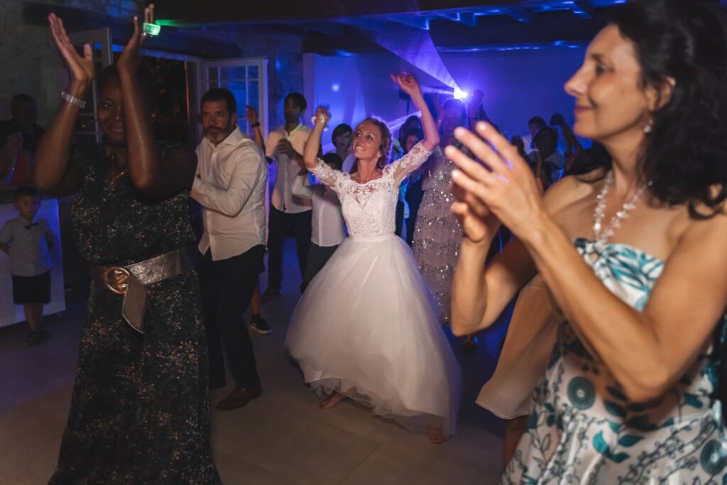 La mariée danse bras levés, entourée d'invités qui applaudissent, sous des lumières colorées.