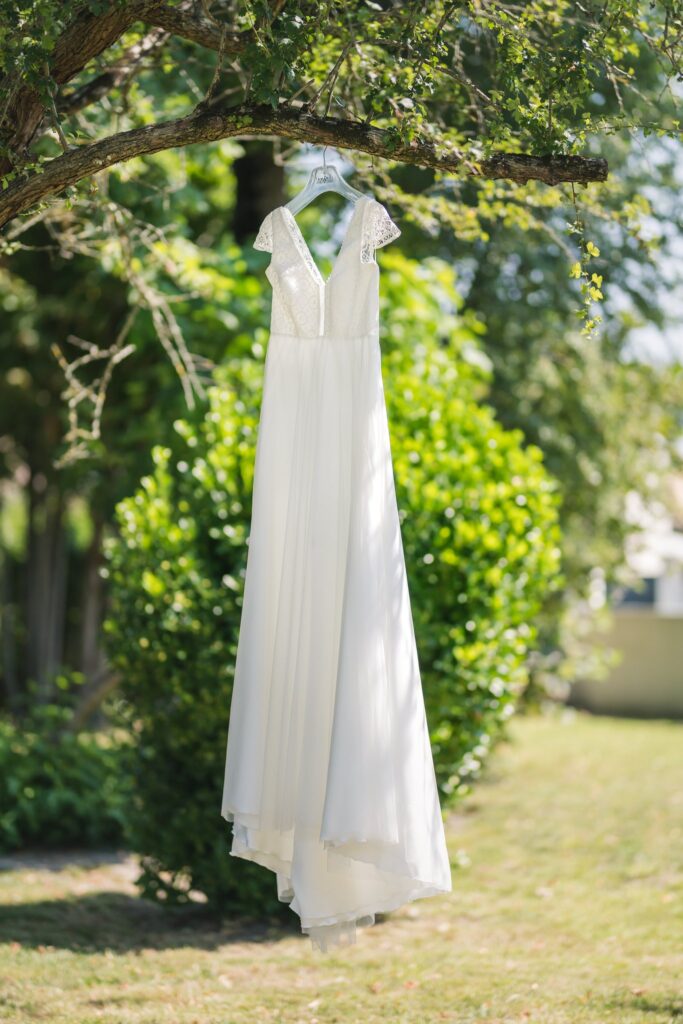 Une robe de mariée blanche est accrochée à une branche d'arbre, devant un buisson et une pelouse ensoleillés.