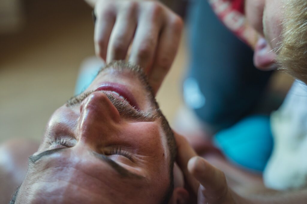 Un homme est allongé sur le dos et se fait raser la barbe. Seuls le visage de l'homme et les mains du barbier sont visibles.