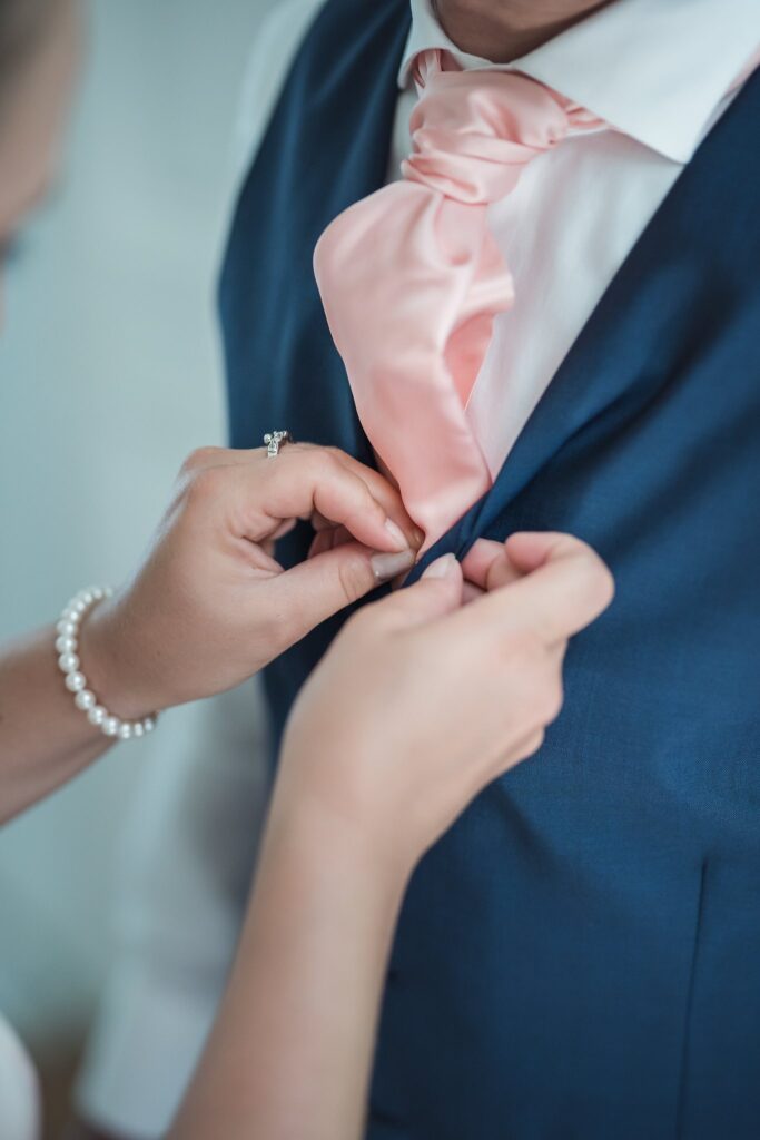 Gros plan des mains d'une femme qui ajuste le gilet bleu d'un homme, avec une cravate rose. La femme porte un bracelet de perles.