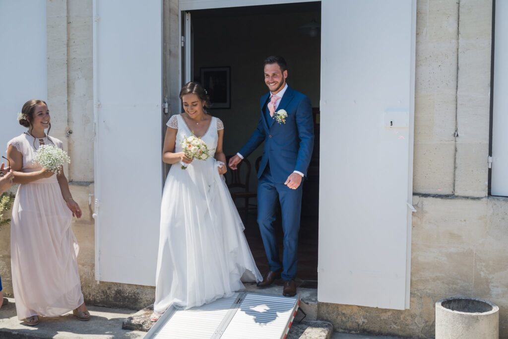 La mariée en robe blanche et le marié en costume bleu sortent par la porte ouverte d'une église en pierre, suivis d'une femme en robe rose pâle.