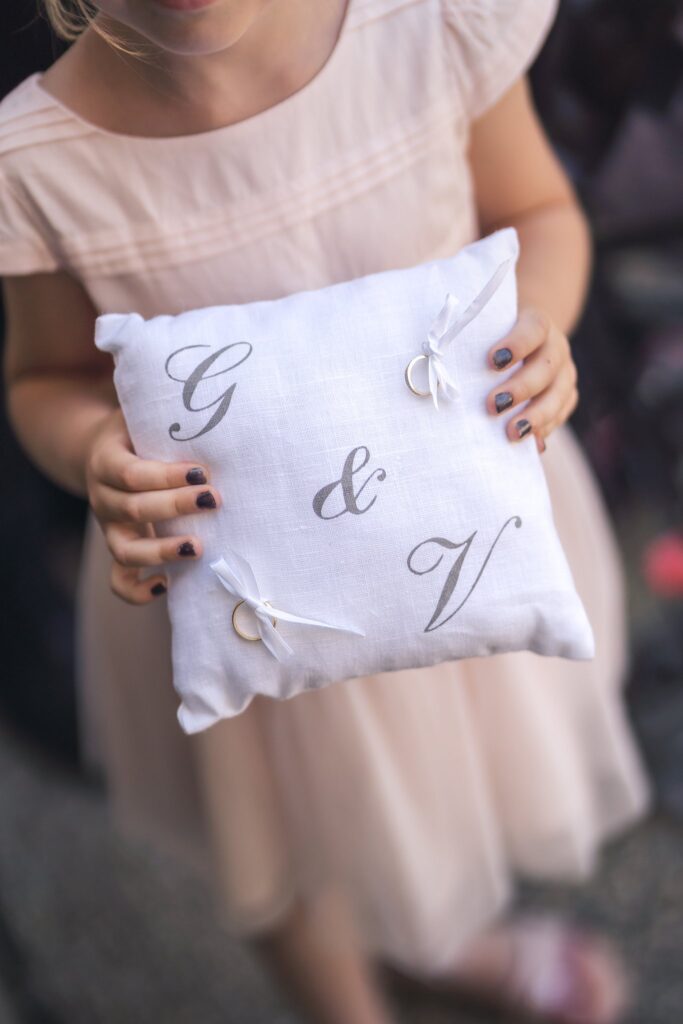 Une fillette en robe rose tient un coussin blanc brodé des lettres « G & V », avec deux alliances.
