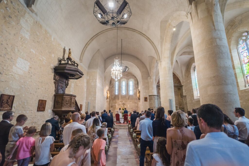 Vue large de l'intérieur d'une église en pierre : les invités sont assis dans la nef, tournés vers le chœur, sous un lustre.