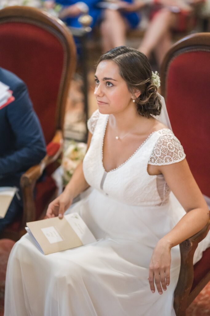 Une femme en robe blanche et voile est assise sur une chaise rouge dans une église. Elle tient un livret à la main.