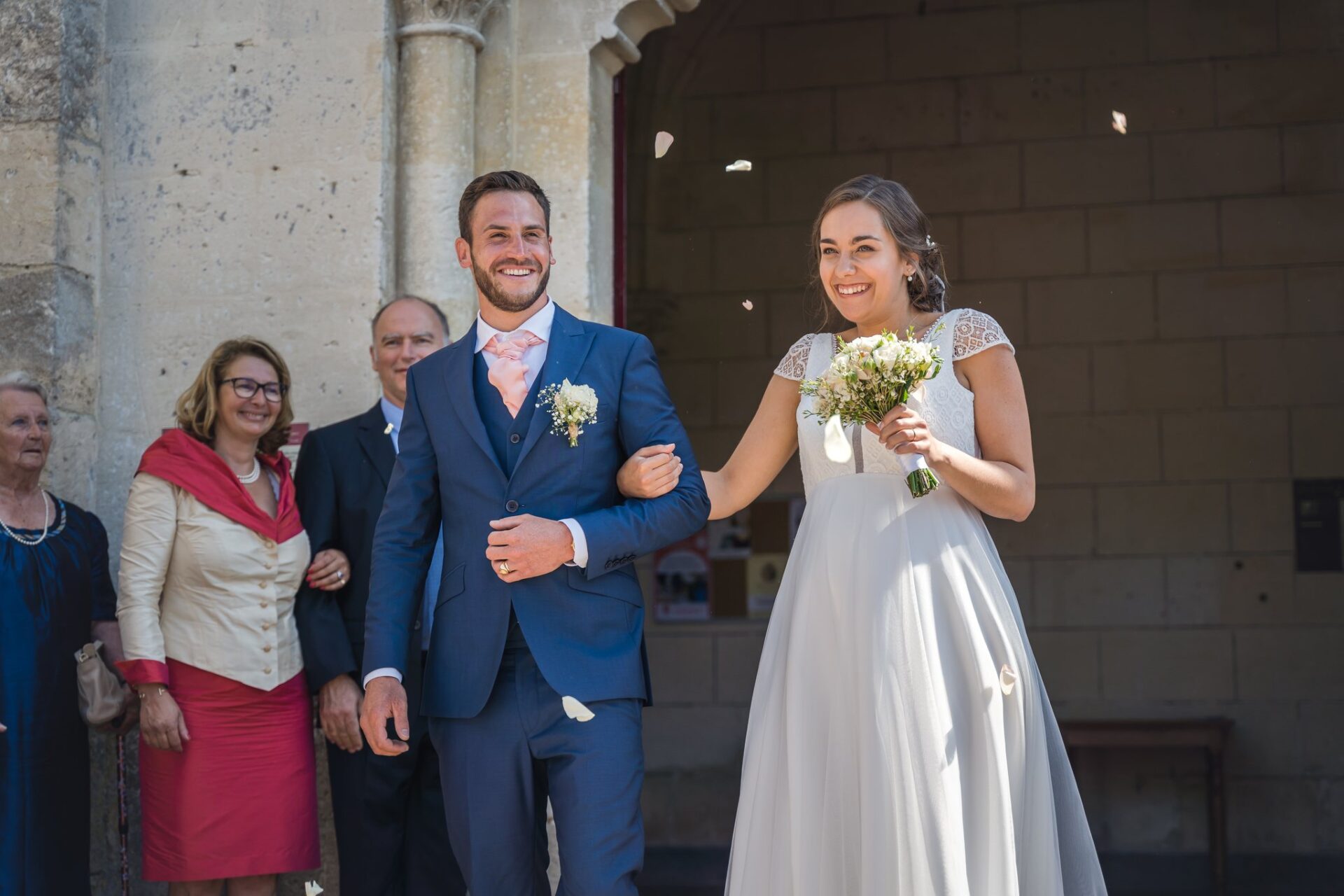Les mariés sortent de l'église bras dessus bras dessous, des pétales dans l'air, devant des invités.