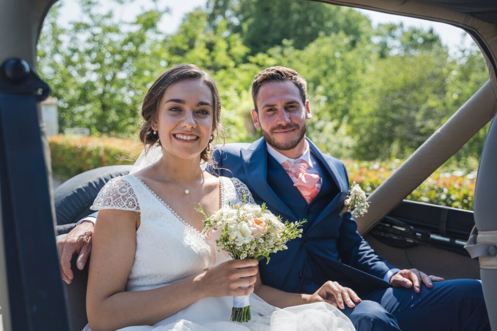 Un couple, la mariée en blanc et le marié en bleu, sourit à l'objectif depuis le siège arrière d'une jeep ouverte. La mariée tient un bouquet de fleurs.