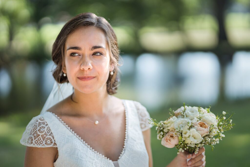 Portrait de la mariée en robe blanche à dentelle, tenant un bouquet, devant un plan d'eau et des arbres flous.