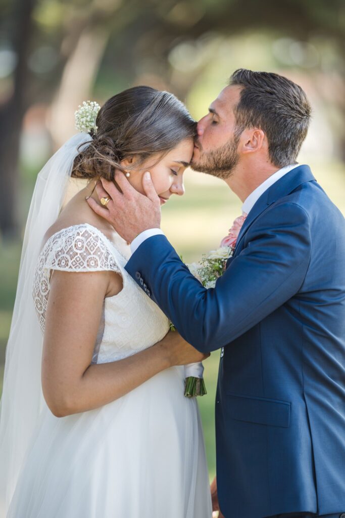 Le marié embrasse le front de la mariée, qui porte une robe blanche et un voile.
