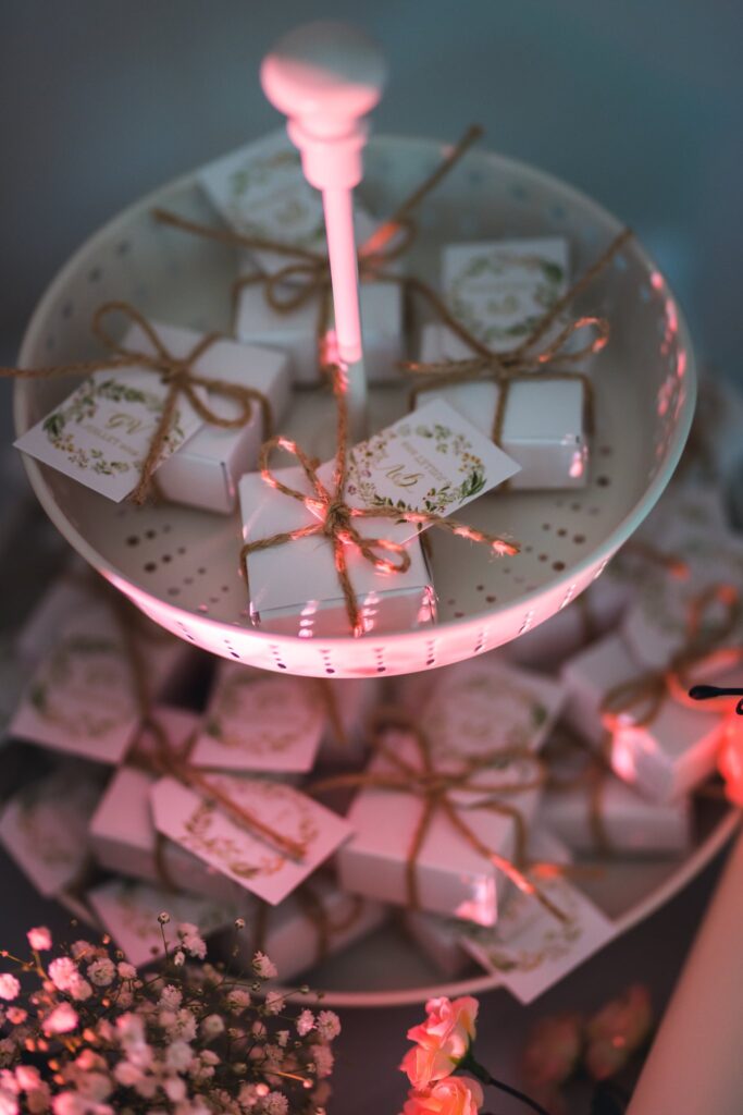 Des petites boîtes blanches nouées de ficelle et ornées d'étiquettes sont présentées sur un présentoir à gâteaux, dans une lumière rose.