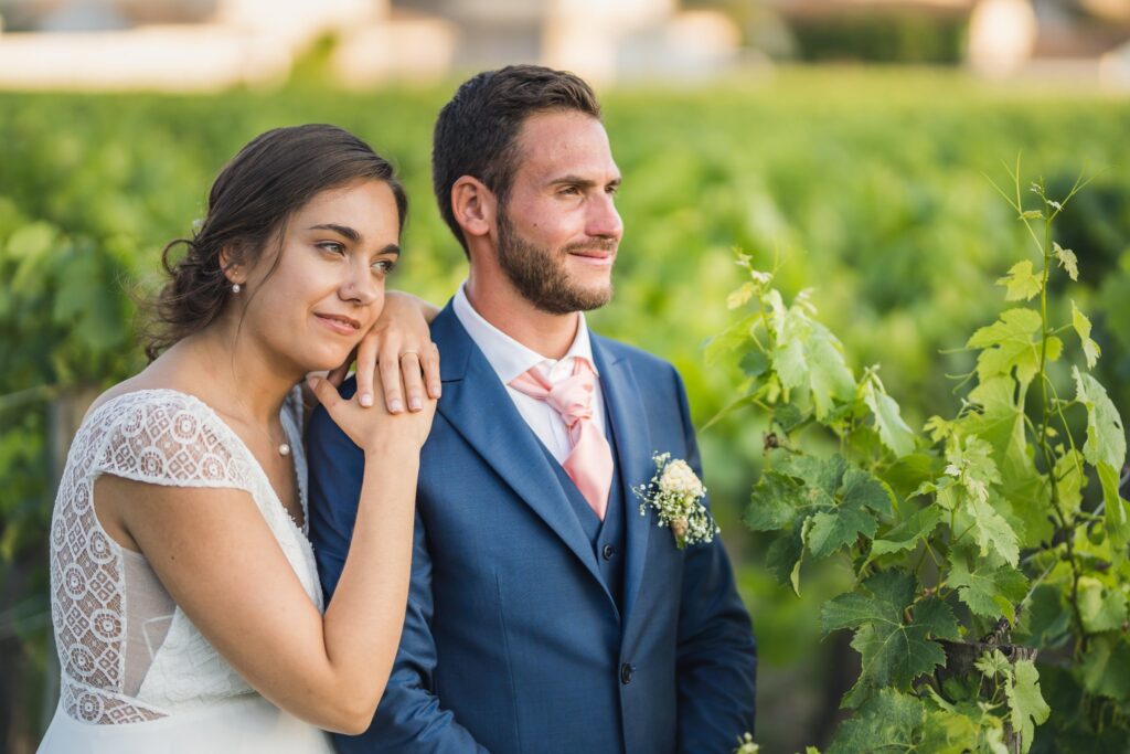Une mariée en robe blanche appuie sa joue sur l'épaule d'un marié en costume bleu, debout dans un vignoble. Le marié regarde au loin.