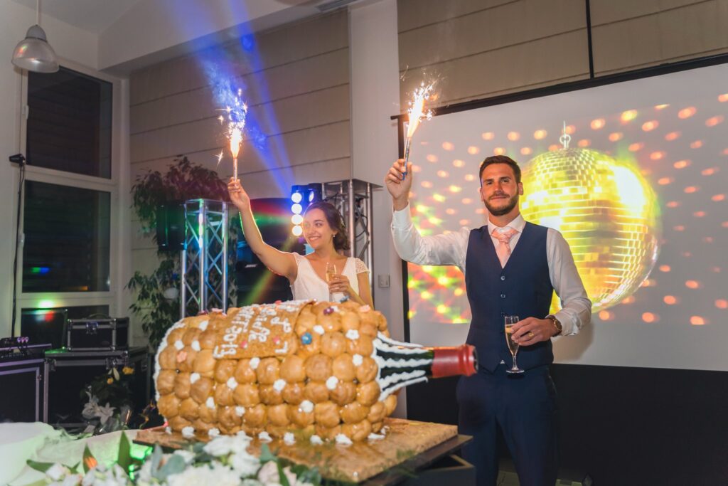 Les mariés souriants tiennent des cierges magiques allumés devant une pièce montée en forme de bouteille de champagne. Une boule disco est visible sur l'écran à l'arrière-plan.