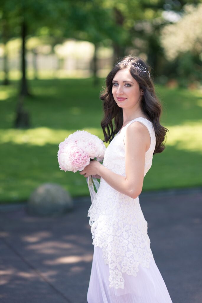 La mariée en robe blanche tient un bouquet de pivoines roses sur une pelouse ombragée.