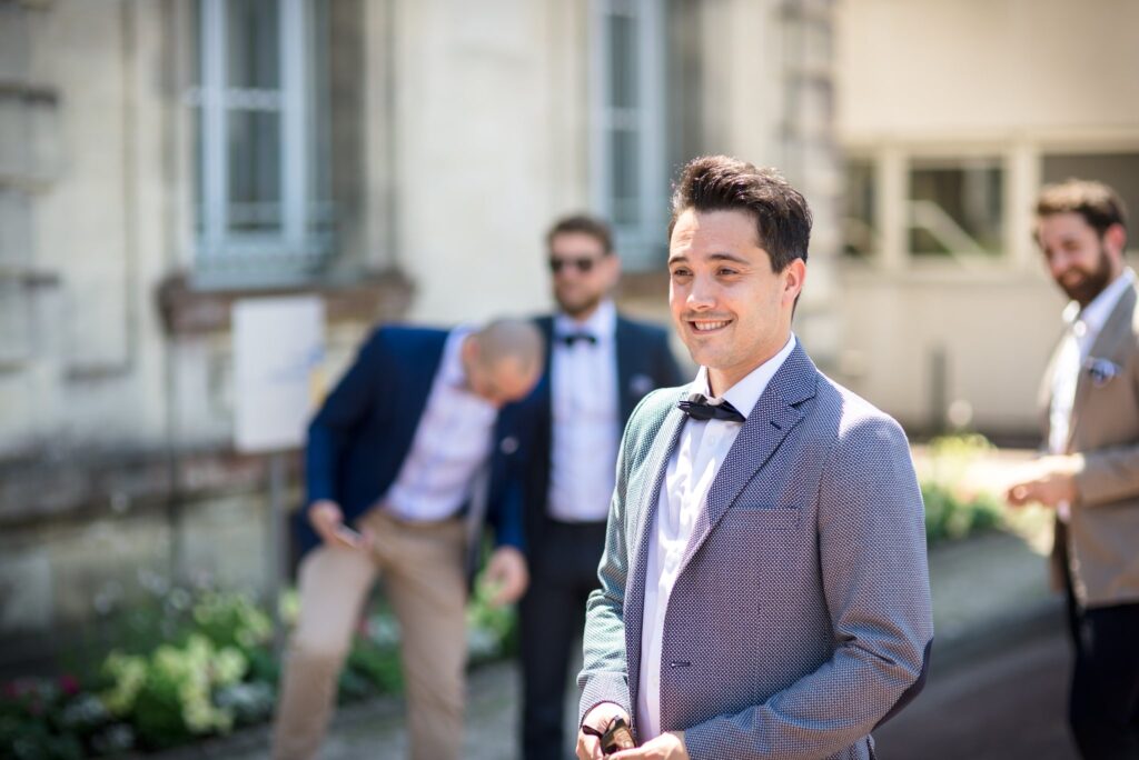 Un homme en veste grise et nœud papillon sourit devant la façade en pierre de la mairie, entouré d'autres invités.