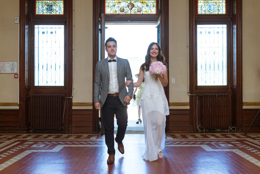 Les mariés entrent côte à côte dans le hall boisé de la mairie, devant de hautes portes vitrées.