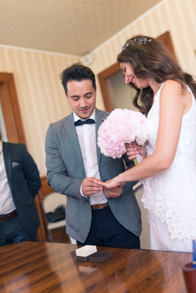 Le marié passe l'alliance au doigt de la mariée, qui tient son bouquet rose.