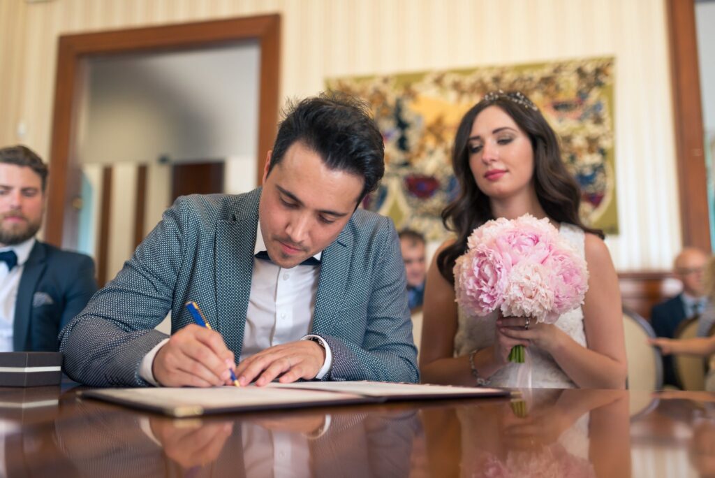 Le marié signe le registre à la table, la mariée à ses côtés avec son bouquet.