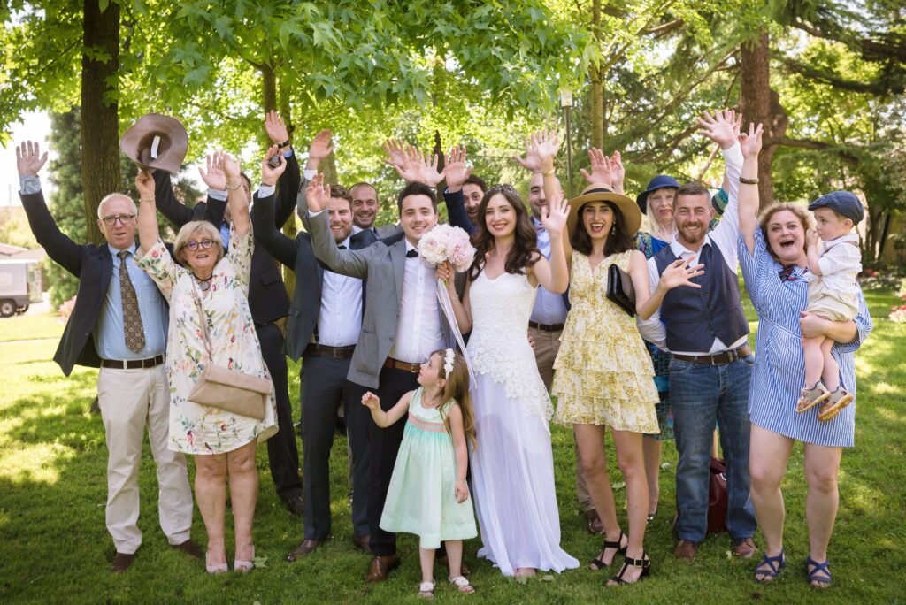 Photo de groupe sur la pelouse : les invités et les mariés lèvent les bras en riant sous les arbres.