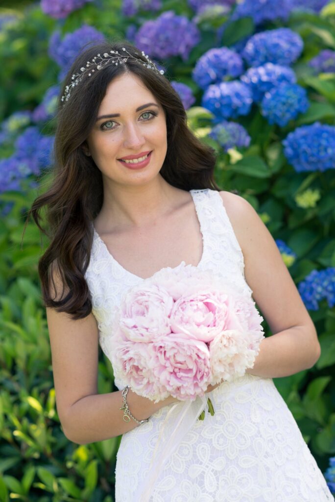 La mariée tient son bouquet rose devant un massif d'hortensias bleus.