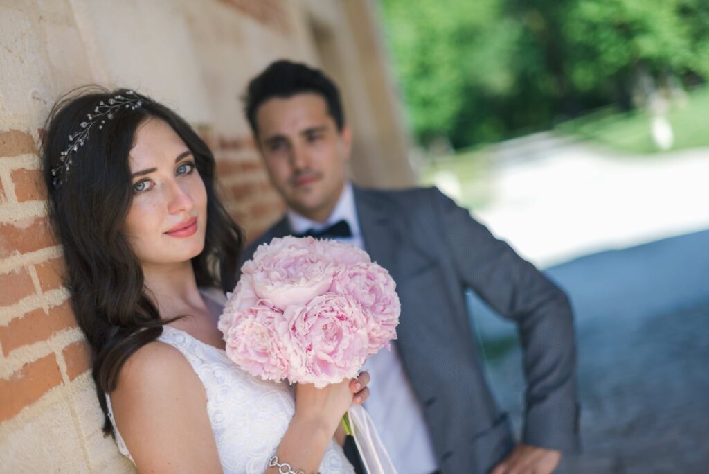 La mariée et le marié posent contre un mur de briques, elle avec son bouquet rose.