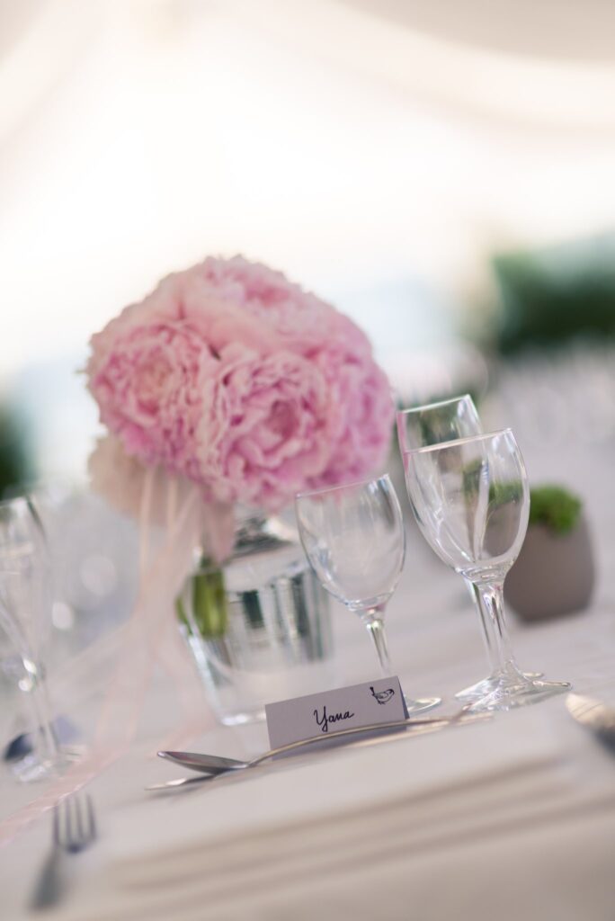 Une pivoine rose dans un petit vase sur une table dressée, avec des verres en cristal.