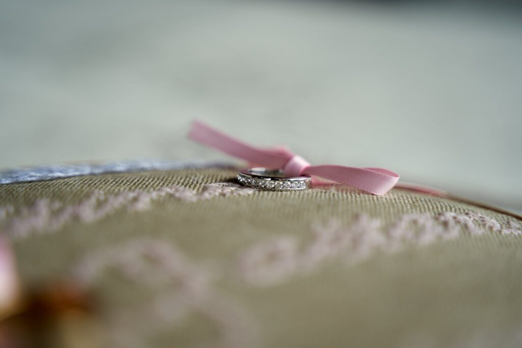 Gros plan sur deux alliances en diamants posées sur un coussin beige clair décoré de dentelle. Un fin ruban rose est noué autour des alliances.
