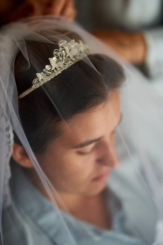 Gros plan sur une femme brune avec un voile blanc et un diadème argenté, dont les mains d'une autre personne ajustent le voile. La femme regarde vers le bas.