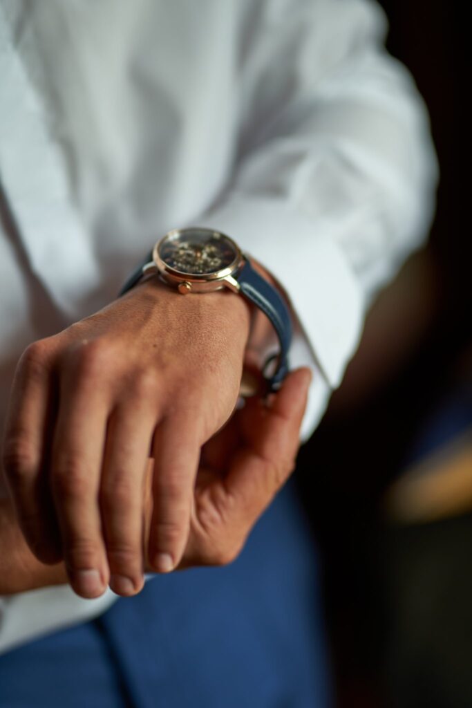Gros plan sur le poignet d'un homme en chemise blanche et veste bleue portant une montre à bracelet bleu. On peut voir le cadran de la montre.