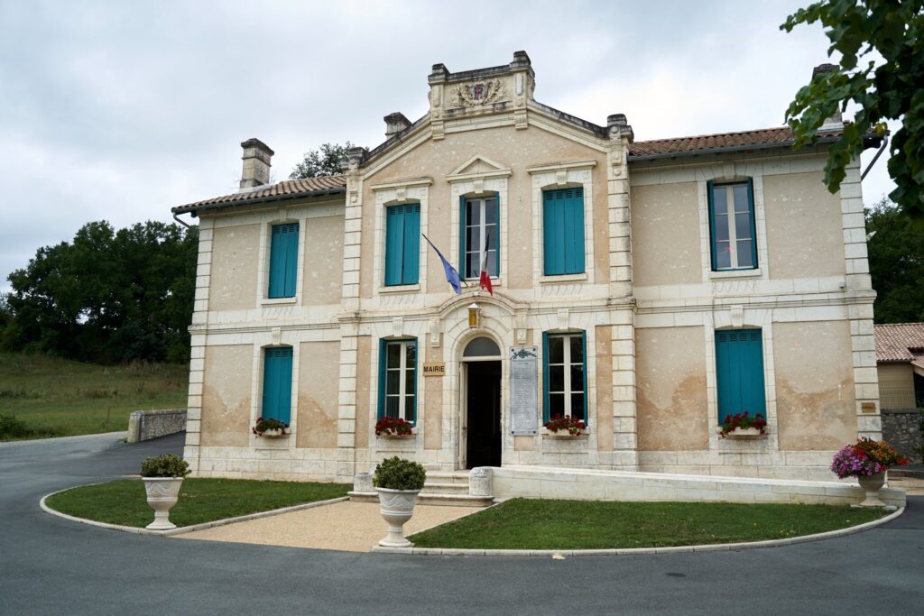 Vue d'ensemble de la façade d'une mairie en pierre claire avec des volets bleus. Des pots de fleurs rouges sont accrochés aux fenêtres et placés devant l'entrée.