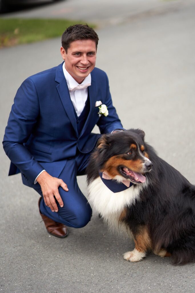 Un homme en costume bleu souriant est accroupi caressant un chien noir et marron avec un nœud papillon. Les deux se trouvent sur une route.