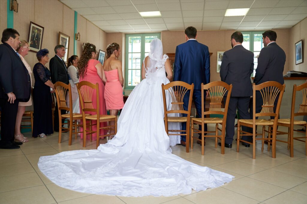 Vue de dos d'une mariée en robe blanche à longue traîne, entourée d'invités dans une salle. Des chaises en bois sont disposées face aux personnes.