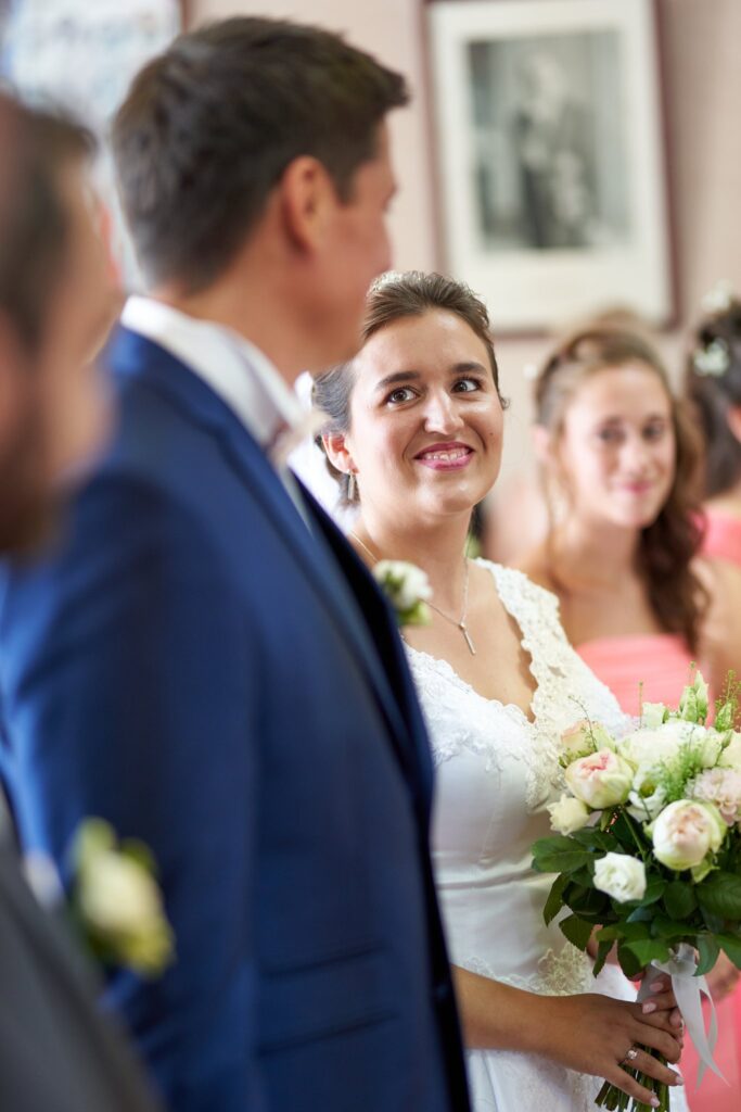 Une mariée en robe blanche et bouquet à la main regarde un homme en costume bleu. Derrière elle, une jeune femme en robe rose.