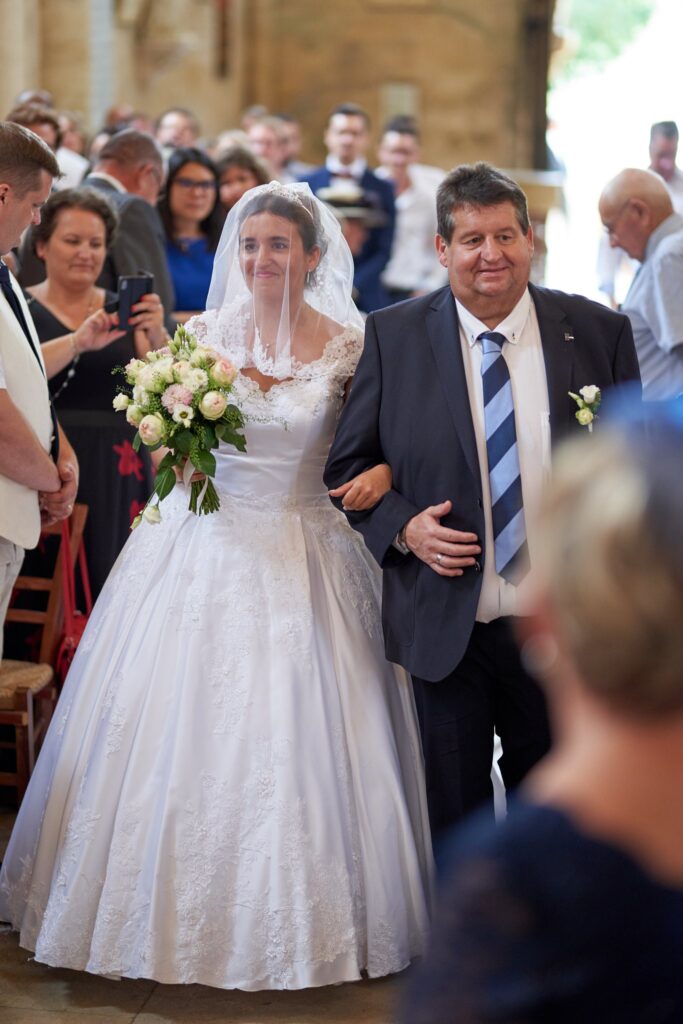 Une mariée en robe blanche et voile, tenant un bouquet, marche à l'église au bras de son père en costume sombre. Des invités sont autour d'eux.