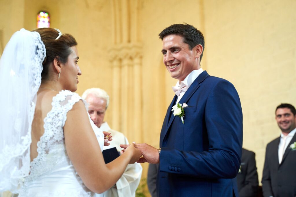 Une mariée en robe blanche place une alliance au doigt d'un marié en costume bleu. Un prêtre est visible derrière eux.