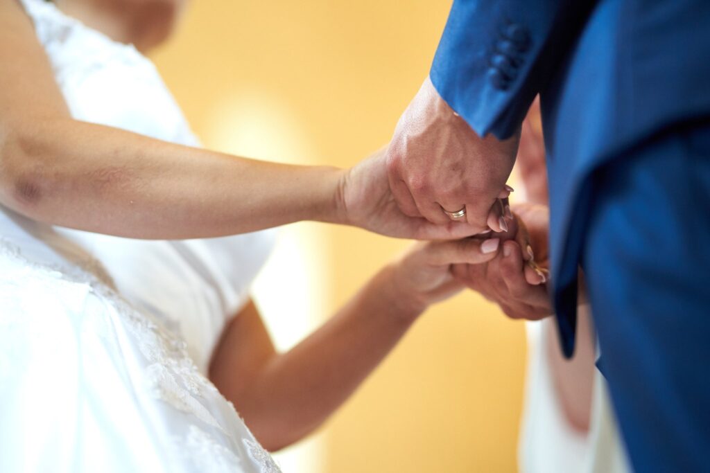 Gros plan des mains entrelacées d'une mariée en robe blanche et d'un marié en costume bleu. On voit une bague dorée à l'annulaire de l'homme.