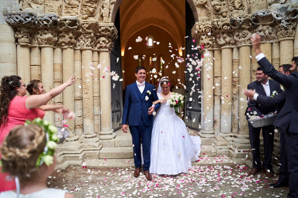 Un couple de mariés en costumes bleu et blanc est à la sortie d'une église en pierre, entourés d'invités qui lancent des pétales de rose.