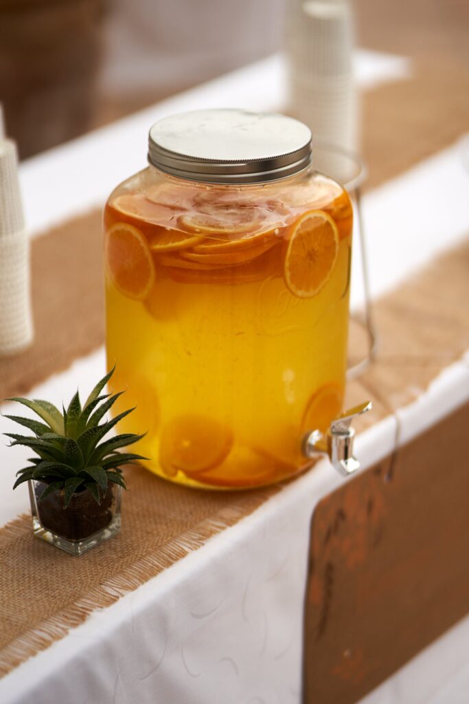 Un grand distributeur de boisson en verre rempli d'une boisson jaune avec des rondelles d'orange. Il est posé sur une table décorée.