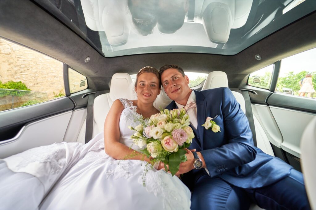 Un couple de mariés souriants est assis et enlacé à l'arrière d'une voiture. La mariée tient son bouquet.