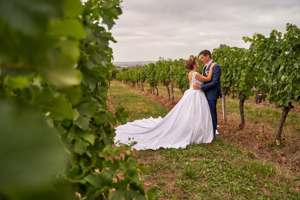 Une mariée en robe blanche et un marié en costume bleu se tiennent enlacés dans un champ de vignes. Ils se regardent avec affection.