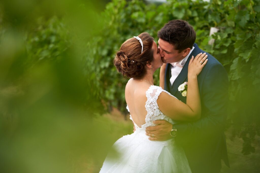 Une mariée en robe blanche et un marié en costume bleu s'embrassent. Le couple est partiellement caché par des feuilles de vigne.