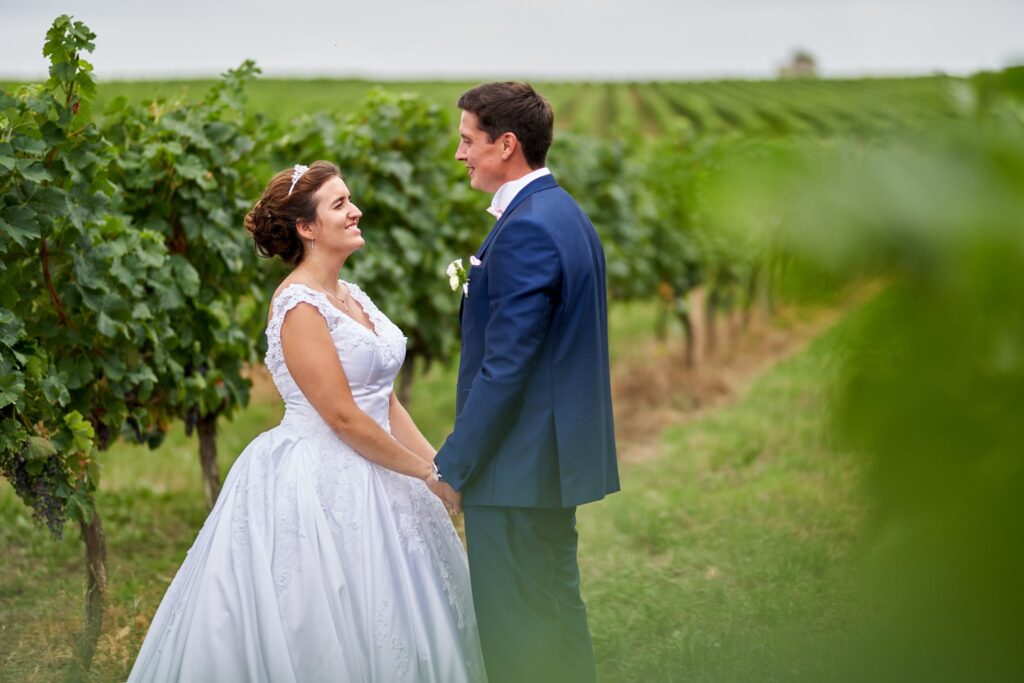Une mariée en robe blanche et un marié en costume bleu se tiennent la main et se regardent dans un champ de vignes. Ils sourient.