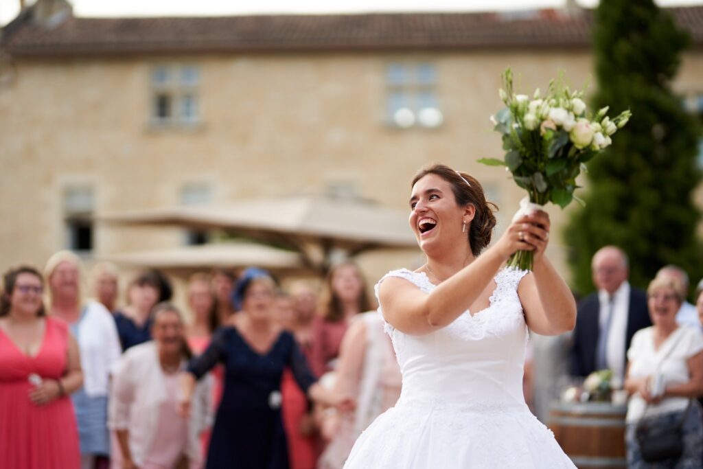 Une mariée en robe blanche, souriante, s'apprête à lancer son bouquet derrière elle. Des femmes et des hommes sont visibles en second plan.