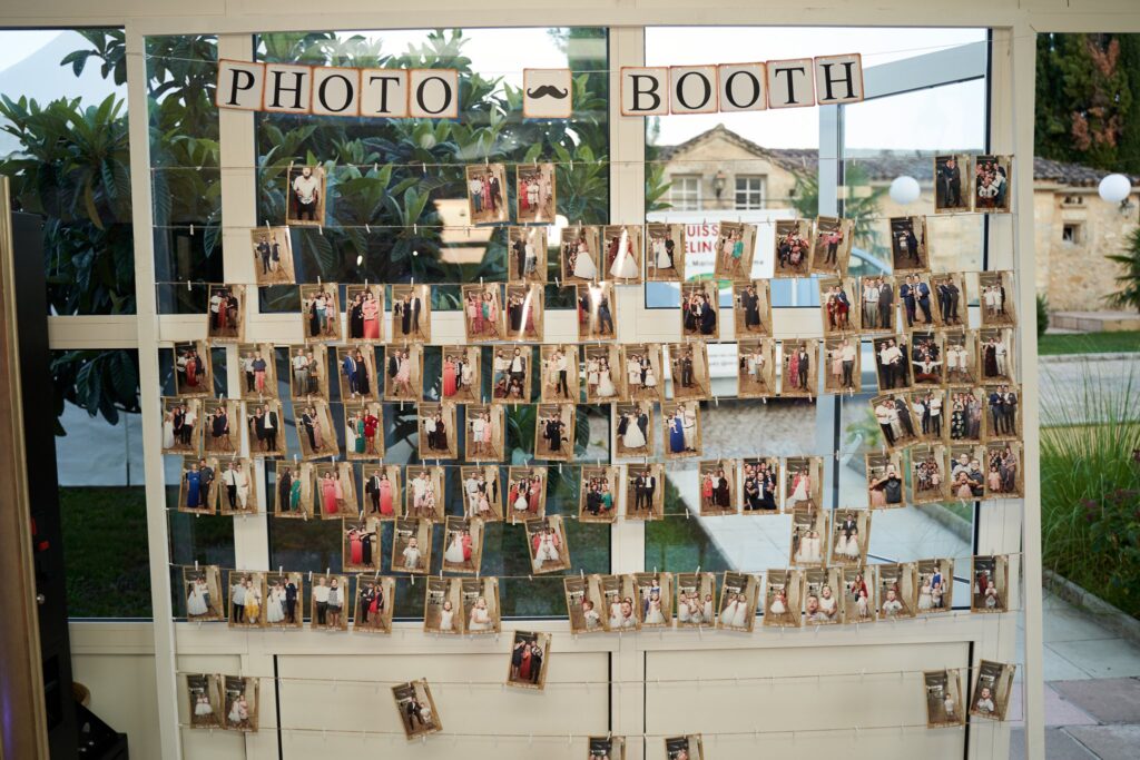 Un panneau décoré avec l'inscription PHOTO BOOTH et des photos accrochées par des pinces à linge. Une maison est visible en arrière-plan.