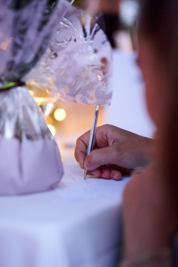 Une personne, vue de dos, signe un livre ou un registre avec un stylo argenté. Un cadeau joliment emballé et des lumières sont visibles en arrière-plan.