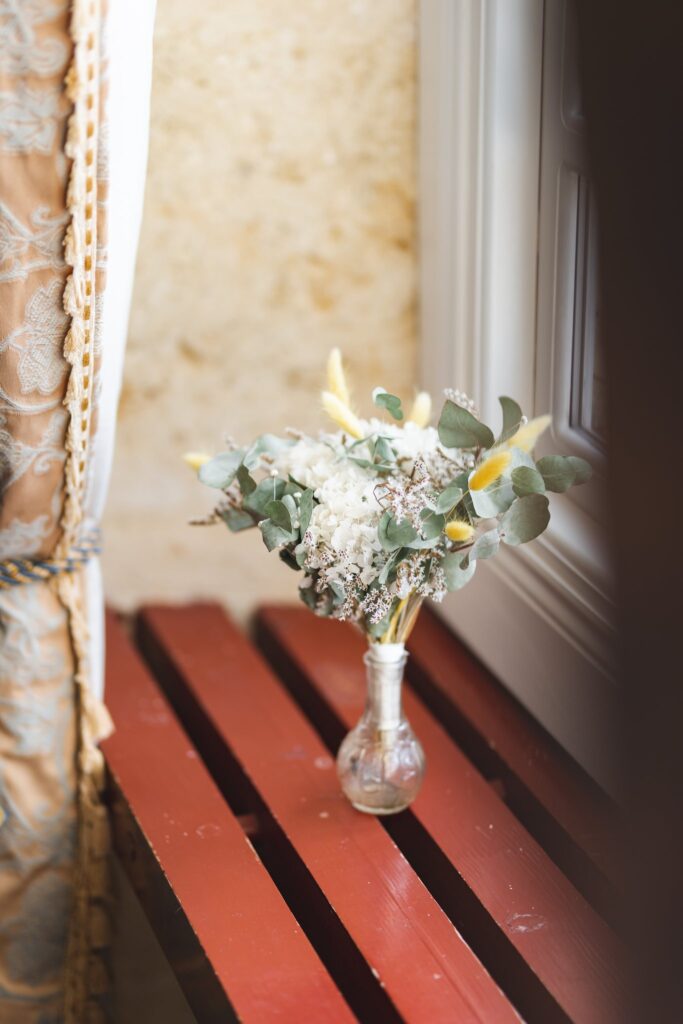 Un petit bouquet de fleurs dans un vase posé sur un banc rouge, devant une fenêtre.