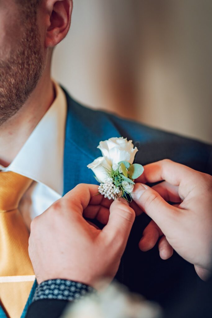 Des mains ajustent une boutonnière de roses blanches sur la veste bleue d'un marié.