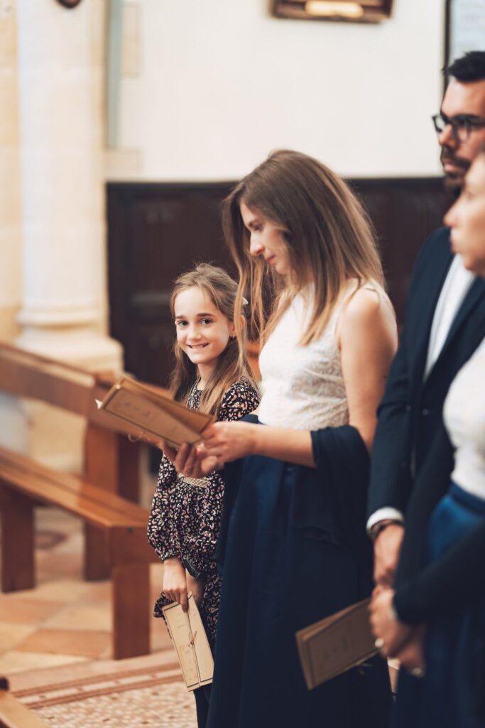 Des invités debout dans les bancs d'une église, dont une fillette qui sourit, tiennent un livret.