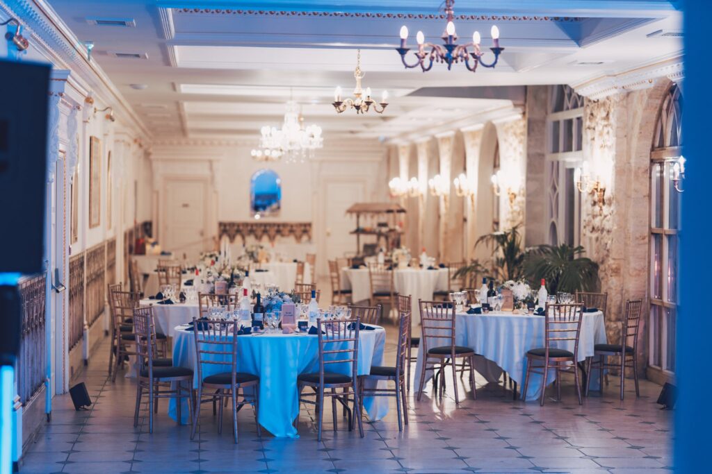 Une grande salle de réception avec lustres et tables rondes dressées en bleu.