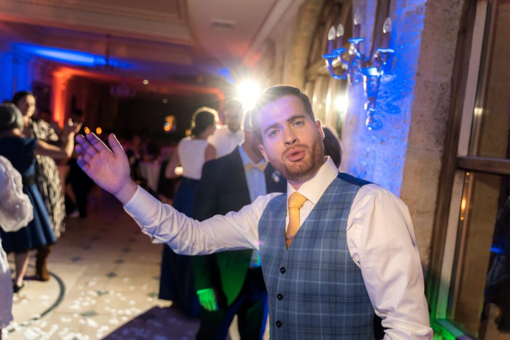 Un homme en gilet à carreaux lève un bras sur la piste de danse, sous des lumières colorées.