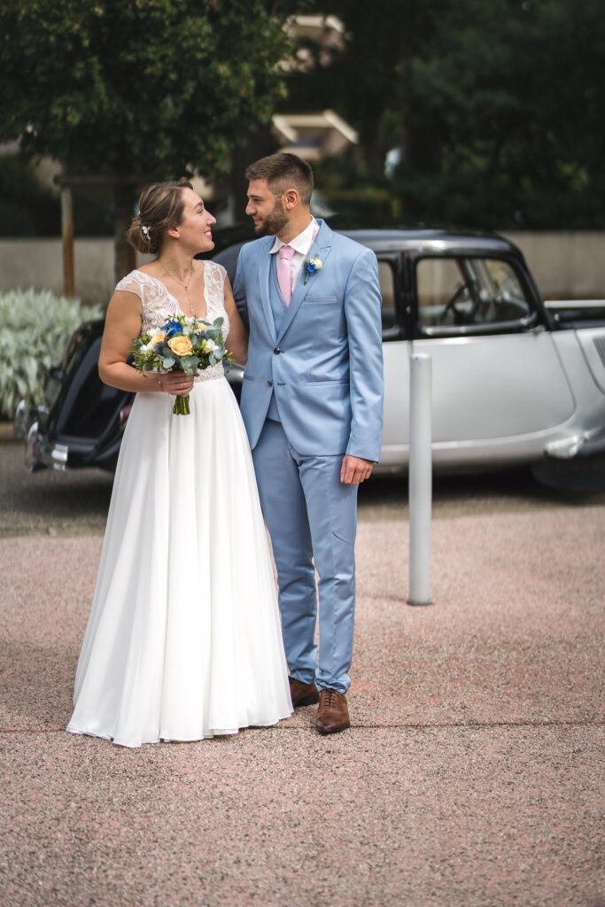Une mariée en robe blanche tenant son bouquet et un marié en costume bleu clair, debout devant une voiture ancienne noire.